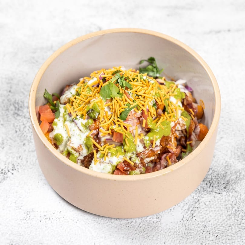 A bowl of Indian chaat with green and white sauces, tomatoes, and crispy yellow noodles.