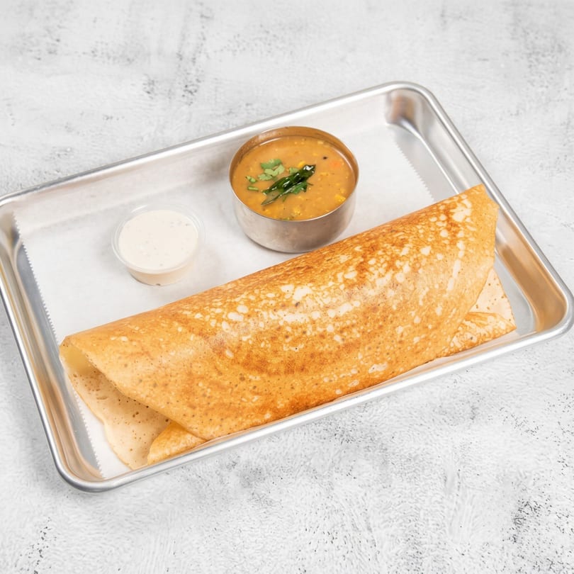 A golden-brown dosa with sambar and chutney on a metal tray with white parchment paper.